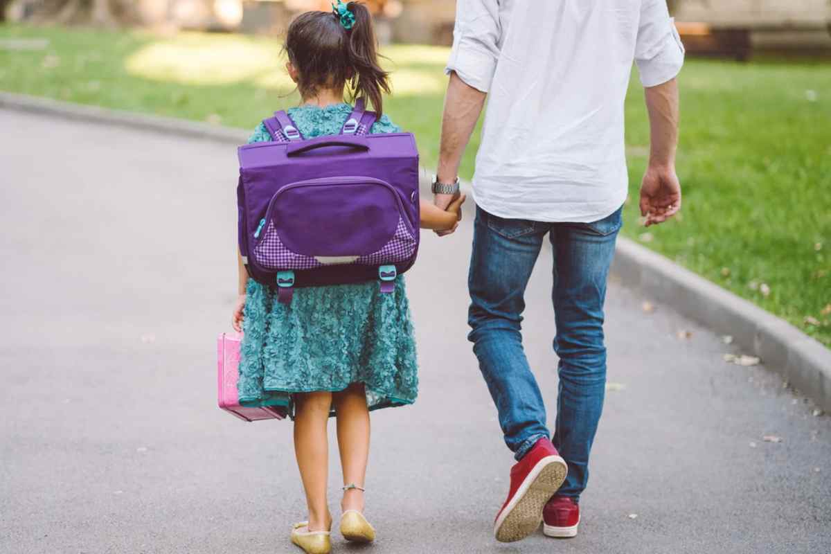 Una bambina con lo zaino viola che tiene per mano il papà